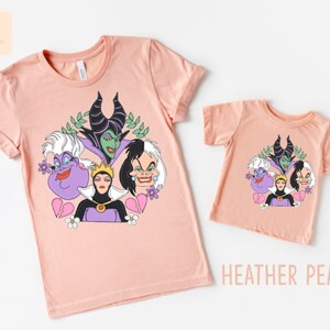 Puede incluir: Dos camisetas de color melocot&oacute;n con un estampado gr&aacute;fico de cuatro personajes de villanos de Disney: &Uacute;rsula, Mal&eacute;fica, la Reina Malvada y Cruella de Vil. El texto "HEATHER PEACH" est&aacute; impreso debajo de la camiseta m&aacute;s peque&ntilde;a.