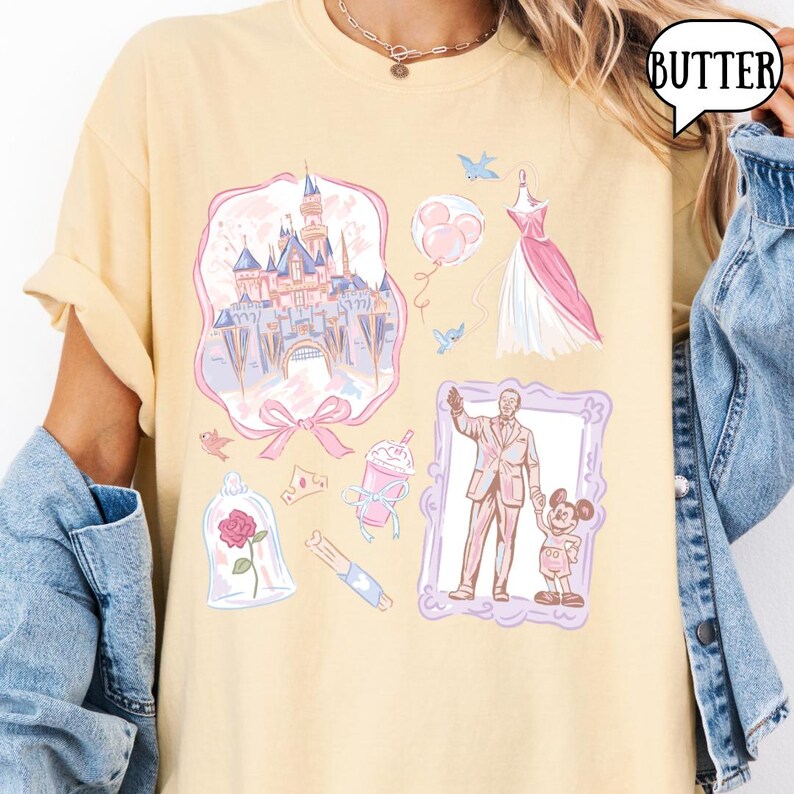 Op de afbeelding: Een lichtgeel T-shirt met diverse Disney-thema illustraties in roze en blauw. Het ontwerp bevat een kasteel, jurk, ballonnen en een ingelijste afbeelding van een man en Mickey Mouse. Het woord "BUTTER" staat in een tekstballon.