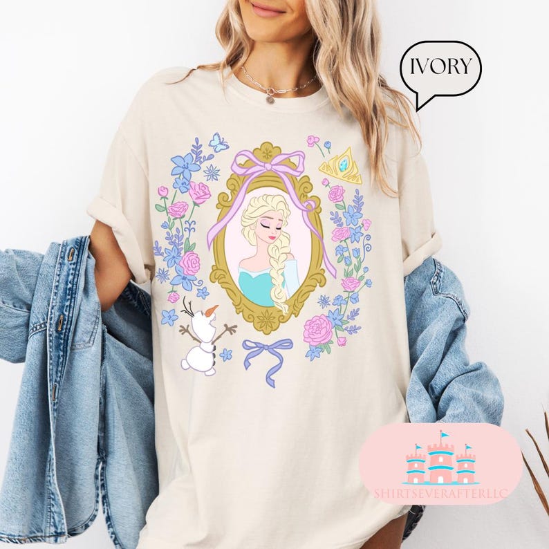 K&ouml;nnte beinhalten: Ein hellbeiges T-Shirt mit einem floralen Design, das ein gerahmtes Bild von Elsa aus Frozen zeigt. Das Design enth&auml;lt auch eine Schneeflocke, eine Krone und eine Schleife. Der Text "IVORY" steht in einer Sprechblase in der oberen rechten Ecke des Bildes.