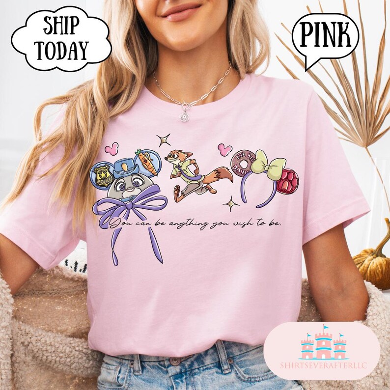 May include: Pink t-shirt featuring cartoon characters and the text "You can be anything you wish to be." The design includes a bunny in a police hat, a fox, and a donut headband. The shirt also has the text "PINK" in a speech bubble.