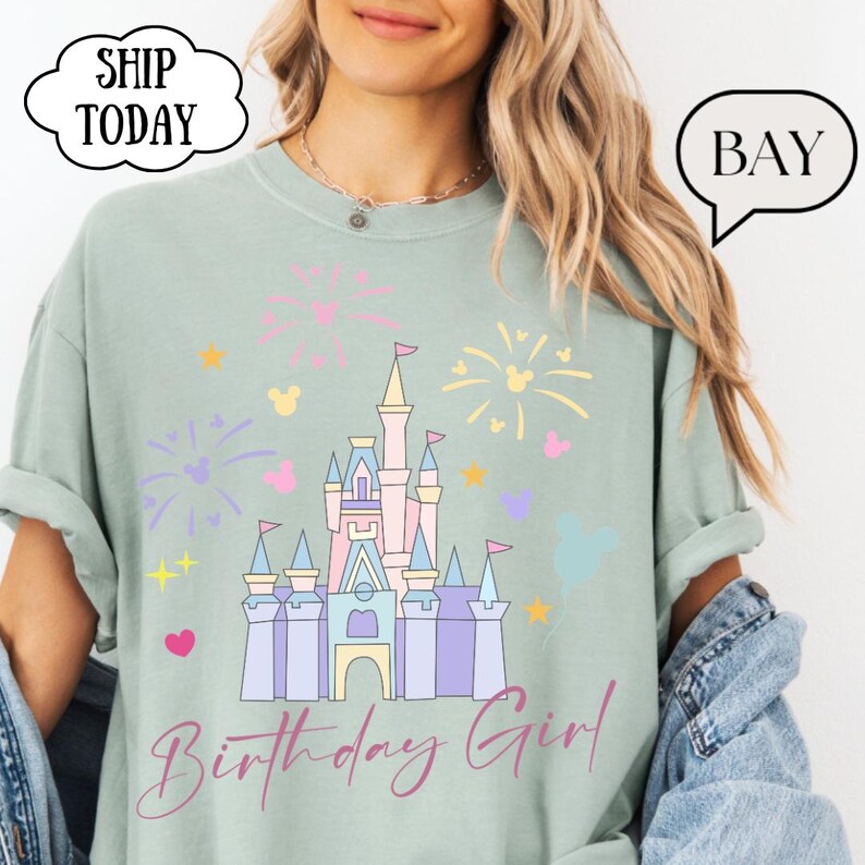 May include: A pale green t-shirt with a castle graphic, fireworks, and Mickey Mouse shapes. The text "Birthday Girl" is in cursive. The shirt is a relaxed fit, suitable for casual wear.