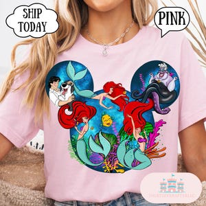 Puede incluir: Una camiseta rosa claro con un dise&ntilde;o tem&aacute;tico de Disney. El dise&ntilde;o presenta personajes de La Sirenita, incluyendo a Ariel y &Uacute;rsula, dispuestos en forma de orejas de Mickey Mouse. La palabra "PINK" est&aacute; en un bocadillo.