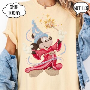 May include: A pale yellow t-shirt with an embroidered design of Mickey Mouse as a wizard. Mickey is wearing a blue hat with a crescent moon, a red robe, and holding a wand. The text "SHIP TODAY" is in a cloud-shaped bubble.