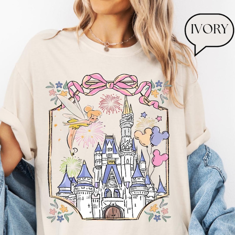Puede incluir: Camiseta color marfil con una ilustraci&oacute;n de castillo de cuento de hadas, un lazo rosa, fuegos artificiales y Campanilla. El dise&ntilde;o incorpora globos pastel y detalles florales, creando un tema m&aacute;gico y festivo.