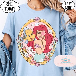 May include: Baby blue t-shirt featuring a colorful illustration of Ariel from The Little Mermaid, holding a pink starfish with Flounder. The design is framed by a gold border with pink seashells and flowers. Text on the shirt says "SHIP TODAY" and "BABY BLUE".