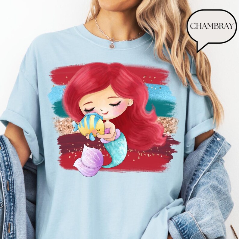 Puede incluir: Camiseta azul claro con un dise&ntilde;o de sirena de dibujos animados. La sirena tiene el pelo rojo, una cola turquesa y sostiene un pez amarillo y azul. El gr&aacute;fico est&aacute; sobre pinceladas rojas, azules y doradas. La palabra "CHAMBRAY" est&aacute; en un bocadillo.
