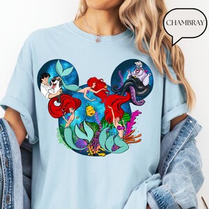 Puede incluir: Camiseta azul claro con un gr&aacute;fico colorido de sirenas y personajes de una pel&iacute;cula de animaci&oacute;n popular. El dise&ntilde;o est&aacute; dispuesto en forma de cabeza de Mickey Mouse. La palabra "CHAMBRAY" est&aacute; en un bocadillo.