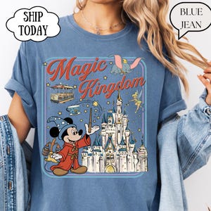 May include: Blue t-shirt featuring a colorful graphic of Mickey Mouse as a sorcerer, a castle, and the words "Magic Kingdom." The design includes Dumbo, Tinkerbell, and a trolley car. The shirt has rolled-up sleeves.