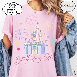 May include: Pale pink t-shirt featuring a castle illustration, fireworks, and the words "Birthday Girl". The design incorporates hearts, stars, and Mickey Mouse shapes. The shirt has short sleeves.