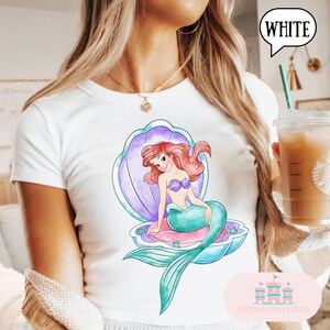 May include: White t-shirt with a watercolour illustration of a mermaid in a seashell. The mermaid has red hair, a purple top, and a teal tail. The seashell is purple and blue. The text "WHITE" is in a speech bubble.