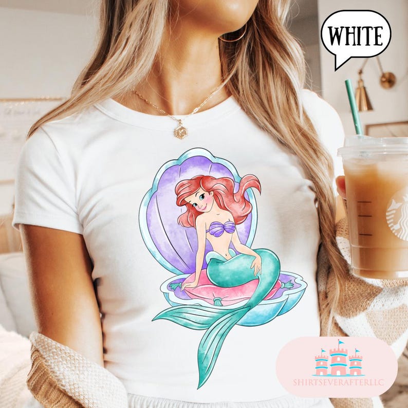 May include: White t-shirt with a watercolour illustration of a mermaid in a seashell. The mermaid has red hair, a purple top, and a teal tail. The seashell is purple and blue. The text "WHITE" is in a speech bubble.