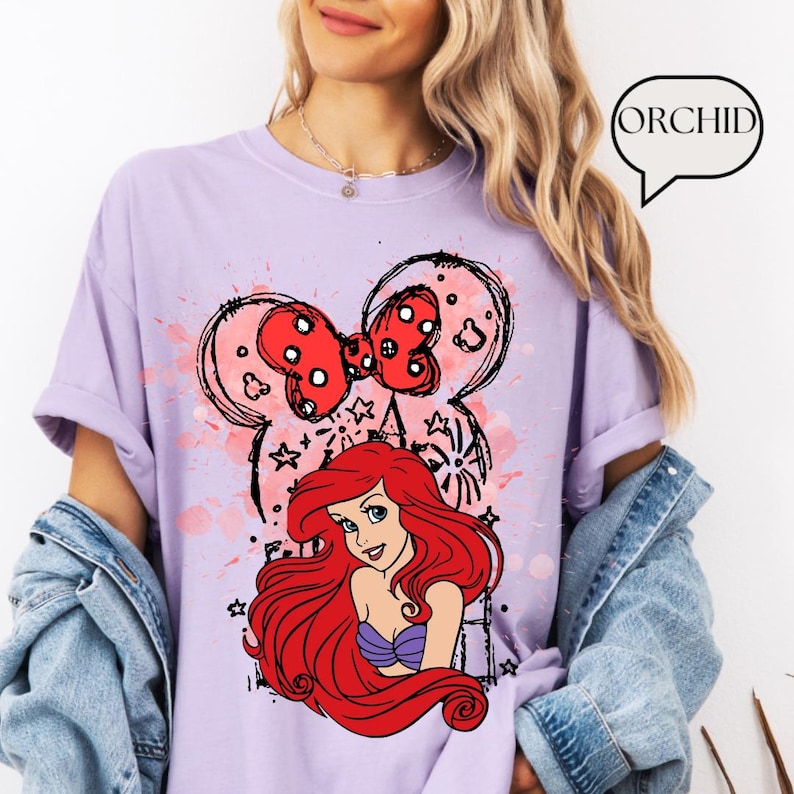 May include: A light purple t-shirt featuring a graphic of Minnie Mouse's head with a red bow and the Little Mermaid, Ariel. The design includes pink paint splatters and black outlines. The word "ORCHID" is in a speech bubble.