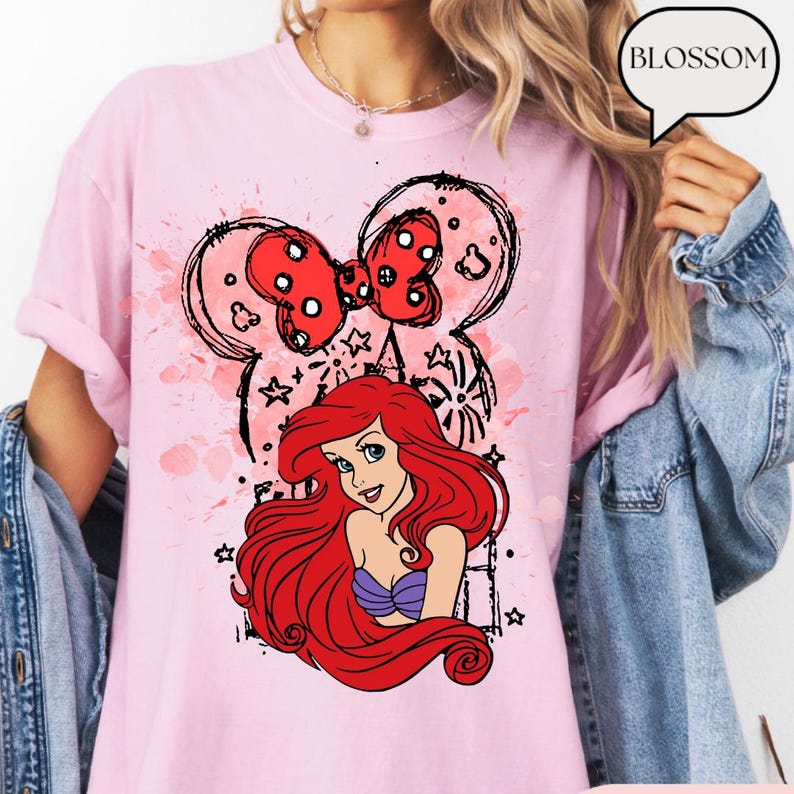 May include: Pink t-shirt featuring a cartoon illustration of Ariel from The Little Mermaid, with a red bow and Mickey Mouse ears design. The shirt has a watercolor-style background and the word "BLOSSOM" in a speech bubble.