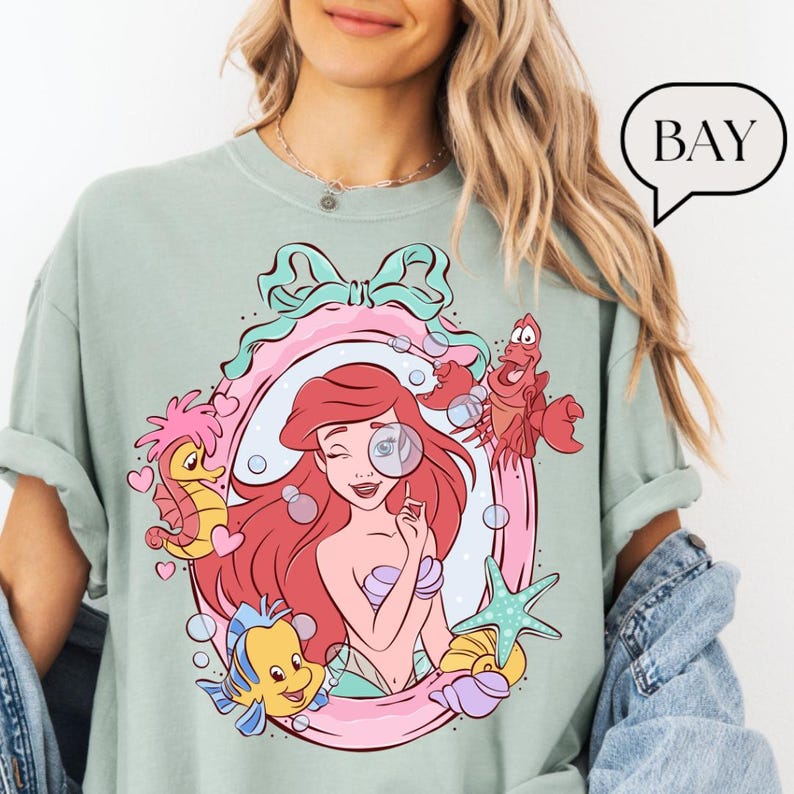 May include: A light green t-shirt featuring a cartoon illustration of Ariel from The Little Mermaid. The design includes Ariel, Flounder, Sebastian, and other sea-themed elements within a pink and blue frame. The word "BAY" is in a speech bubble.