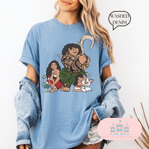 May include: A blue t-shirt with a graphic of Maui, Moana, and Pua from the Disney movie Moana. The shirt is made of washed denim.