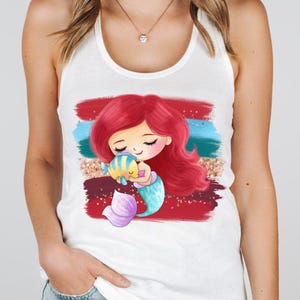 Puede incluir: Camiseta sin mangas blanca con un dise&ntilde;o de sirena de dibujos animados. La sirena tiene pelo rojo, una cola azul y morada, y sostiene un pez amarillo y azul. El fondo presenta pinceladas rojas, azules y doradas.