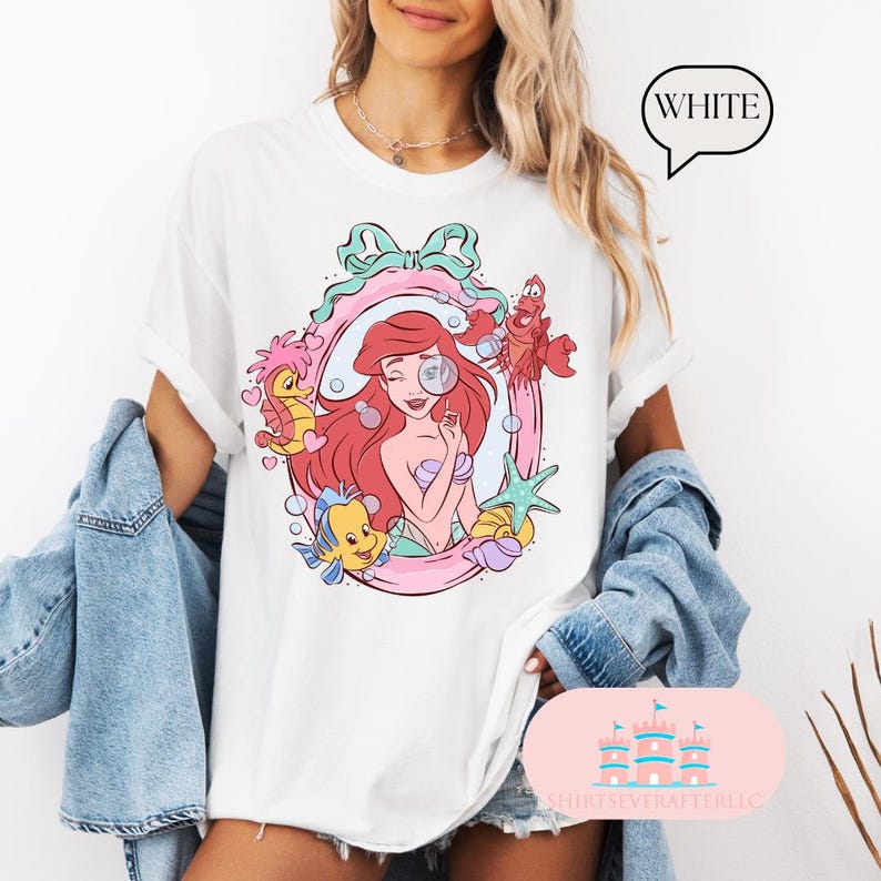 K&ouml;nnte beinhalten: Wei&szlig;es T-Shirt mit einem farbenfrohen Grafik von Arielle, der Meerjungfrau, umrahmt von einem rosa und t&uuml;rkisfarbenen Oval mit Meeresbewohnern. Der Text "WHITE" befindet sich in einer Sprechblase. Das T-Shirt hat hochgekrempelte &Auml;rmel.