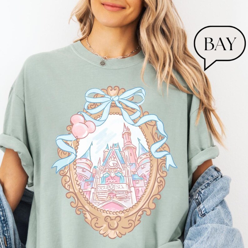 Puede incluir: Camiseta verde claro con un dise&ntilde;o de castillo de cuento de hadas dentro de un marco ovalado adornado. El marco est&aacute; adornado con un lazo azul claro y globos rosas. La palabra "BAY" est&aacute; en una burbuja.