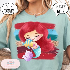 Puede incluir: Camiseta azul polvoriento con un dise&ntilde;o de sirena de dibujos animados. La sirena tiene el pelo rojo y una cola turquesa, abrazando a un pez amarillo y azul. El dise&ntilde;o incluye detalles en rojo, azul y dorado. El texto "Dusty Blue" es visible.