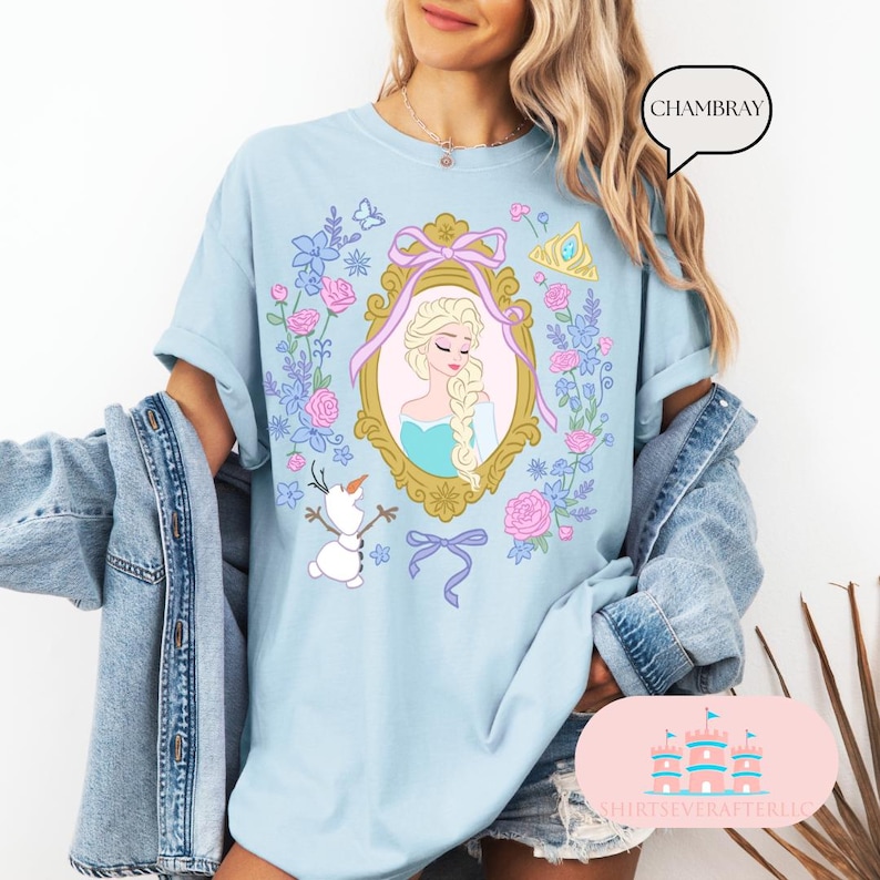K&ouml;nnte beinhalten: Hellblaues T-Shirt mit einem Blumendesign, das ein gerahmtes Bild von Elsa aus Frozen zeigt. Der Text "CHAMBRAY" befindet sich in einer Sprechblase in der oberen rechten Ecke des Bildes.