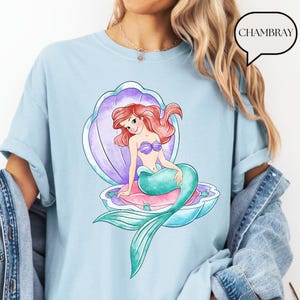 May include: Pale blue t-shirt with a vibrant illustration of a mermaid in a seashell. The mermaid has red hair, a purple seashell bra, and a teal tail. The seashell is purple, pink, and blue. The word "CHAMBRAY" is in a speech bubble.