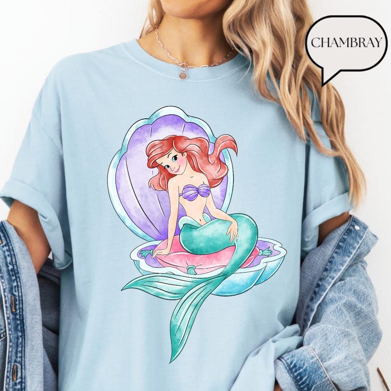 May include: Pale blue t-shirt with a vibrant illustration of a mermaid in a seashell. The mermaid has red hair, a purple seashell bra, and a teal tail. The seashell is purple, pink, and blue. The word "CHAMBRAY" is in a speech bubble.