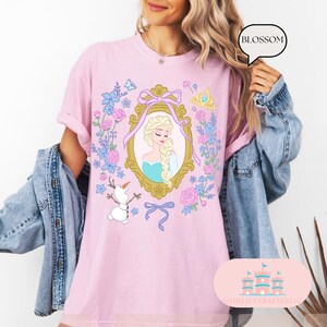K&ouml;nnte beinhalten: Pinkes T-Shirt mit einem floralen Design, das eine Prinzessin in einem goldenen Rahmen, einen Schneemann und das Wort "Blossom" zeigt.