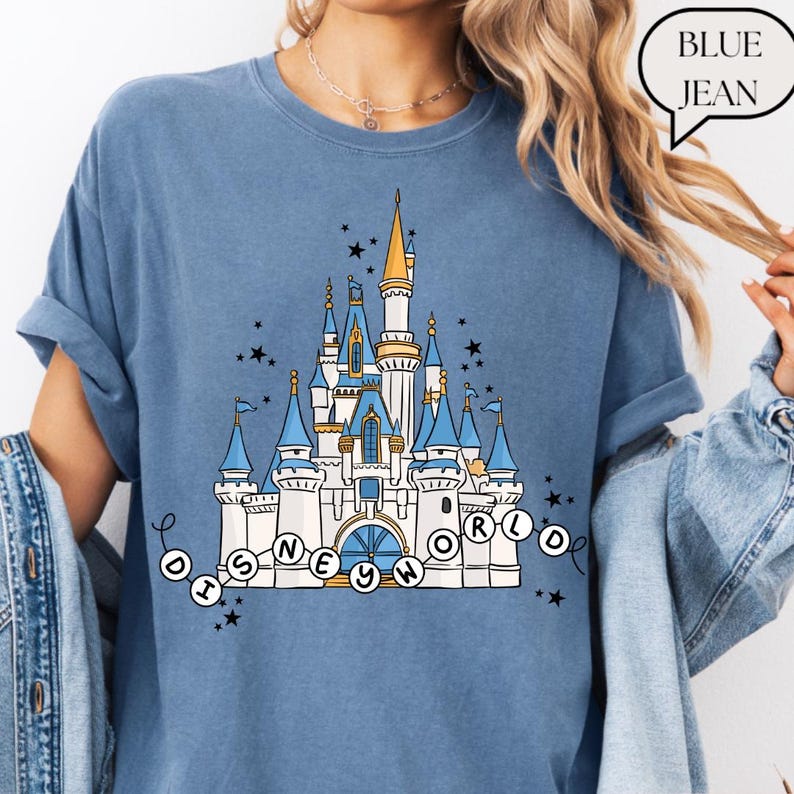 May include: Blue t-shirt featuring a detailed illustration of a fairytale castle with blue turrets, gold accents, and the text "DISNEY WORLD" in a circular design. The shirt is a light blue color.