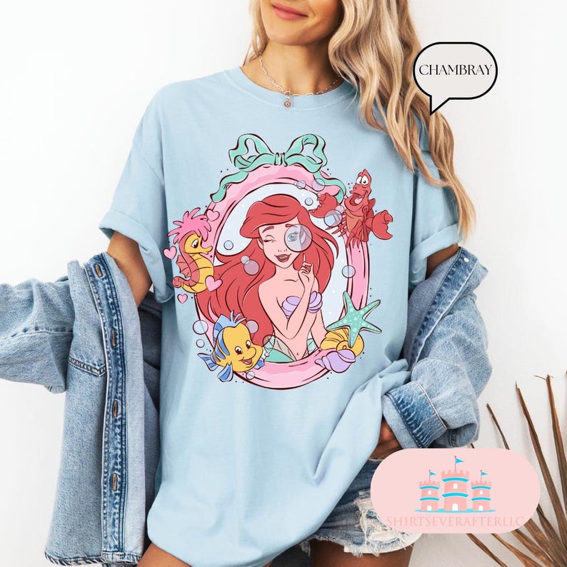 K&ouml;nnte beinhalten: Hellblaues T-Shirt mit einer farbenfrohen Illustration einer Meerjungfrau mit roten Haaren, umgeben von Meeresbewohnern. Das Design umfasst einen rosa und gr&uuml;nen Rahmen, Blasen und den Text "CHAMBRAY". Das Shirt hat eine lockere Passform.