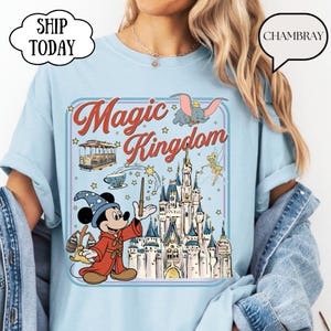 May include: Light blue t-shirt featuring a colorful graphic of Mickey Mouse as a wizard, a castle, and the words "Magic Kingdom." The design includes Dumbo, Tinkerbell, and a cable car. The shirt has a round neck and short sleeves.