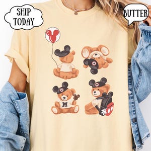 May include: A pale yellow t-shirt featuring a graphic of four brown teddy bears wearing black mouse ear hats. One bear holds a red balloon, one is upside down, one wears a white shirt with an M, and one carries a black and red backpack.