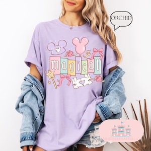Op de afbeelding: Een lichtpaars t-shirt met het woord "magical" in pastelkleuren, met cartoon illustraties van een toverstaf, Mickey Mouse oren, een ballon, strikken, rozen en een konijn. Het shirt heeft korte mouwen en een relaxte pasvorm.