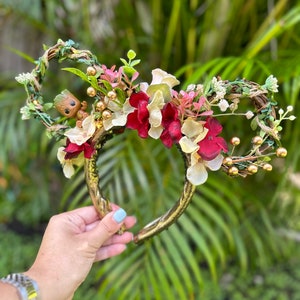 Puede incluir: Diadema dorada con una disposición floral con flores rojas, blancas y verdes. Una pequeña figura marrón de un personaje de Guardianes de la Galaxia está anidada en la vegetación.