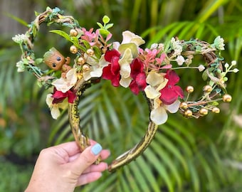 May include: Gold headband with a floral arrangement featuring red, white, and green flowers. A small brown figurine of a character from the Guardians of the Galaxy is nestled in the greenery.