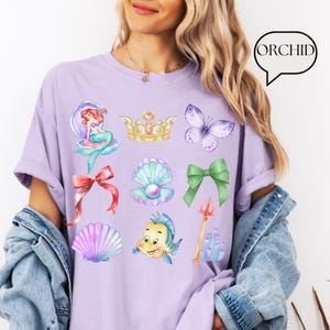 Może przedstawiać: Jasnofioletowa koszulka z kolorowymi ilustracjami syreny, korony, motyla, kokardy, muszli, perły i ryby. Koszulka ma r&oacute;wnież napis "ORCHID" w dymku.