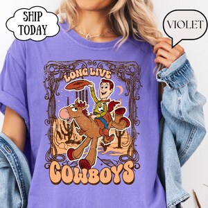 May include: Violet-colored t-shirt featuring a cartoon cowboy riding a horse, with the words "LONG LIVE COWBOYS" in a vintage design. The cowboy is holding a hat. The shirt has a relaxed fit.