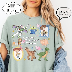 Puede incluir: Una camiseta verde salvia con un estampado gr&aacute;fico de varios personajes de juguete animados, incluyendo un ranger espacial, un vaquero, una vaquera, un dinosaurio, un cerdo, un perro y un alien&iacute;gena. El texto "YOU'VE GOT A FRIEND IN ME" est&aacute; impreso.