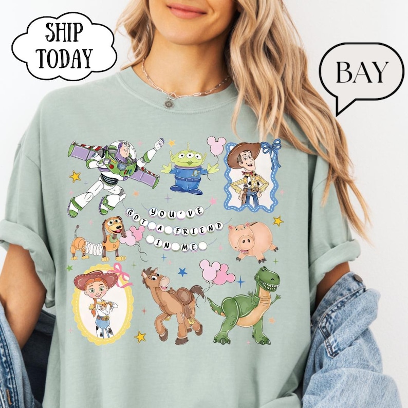 Puede incluir: Una camiseta verde salvia con un estampado gr&aacute;fico de varios personajes de juguete animados, incluyendo un ranger espacial, un vaquero, una vaquera, un dinosaurio, un cerdo, un perro y un alien&iacute;gena. El texto "YOU'VE GOT A FRIEND IN ME" est&aacute; impreso.