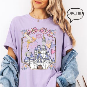 Puede incluir: Una camiseta lila con un castillo, un hada, fuegos artificiales y globos. El gr&aacute;fico est&aacute; enmarcado con un lazo rosa y detalles florales. La palabra "ORCHID" est&aacute; en un bocadillo.