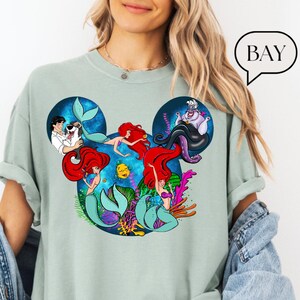 Puede incluir: Camiseta verde claro con un dise&ntilde;o de dibujos animados de personajes de La Sirenita. El dise&ntilde;o incluye a Ariel, el Pr&iacute;ncipe Eric, &Uacute;rsula y Flounder, dispuestos en forma de cabeza de Mickey Mouse. La palabra "BAY" est&aacute; en una burbuja.