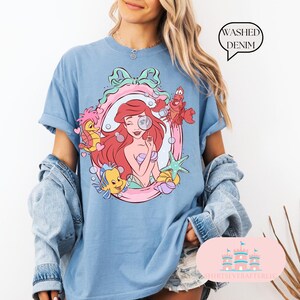 K&ouml;nnte beinhalten: Hellblaues T-Shirt mit einer farbenfrohen Grafik von Arielle, der Meerjungfrau, umgeben von Meereskreaturen. Das Shirt hat hochgekrempelte &Auml;rmel und den Text "Washed Denim" in einer Sprechblase.