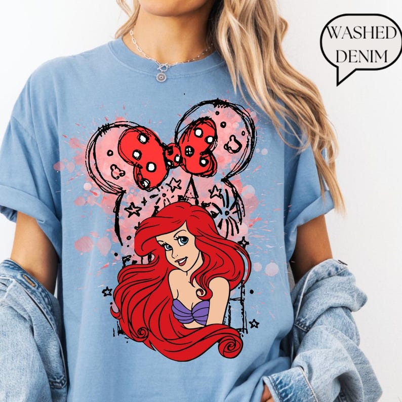 May include: Light blue t-shirt featuring a graphic of Ariel from The Little Mermaid, with a red bow and Mickey Mouse ears design. The shirt has the words "Washed Denim" in a speech bubble.