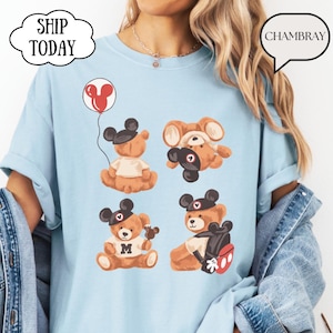 May include: A light blue t-shirt featuring a graphic print of four brown teddy bears wearing black mouse ear hats. One bear holds a red balloon, one is upside down, one wears a shirt with the letter M, and one carries a black backpack.