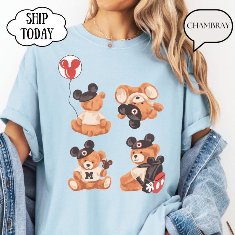 May include: A light blue t-shirt featuring a graphic print of four brown teddy bears wearing black mouse ear hats. One bear holds a red balloon, one is upside down, one wears a shirt with the letter M, and one carries a black backpack.