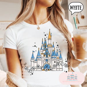 May include: White t-shirt featuring a whimsical illustration of a castle with blue and gold accents, surrounded by stars. The text "DISNEY WORLD" is displayed in a circular design. The shirt is a crew neck style.