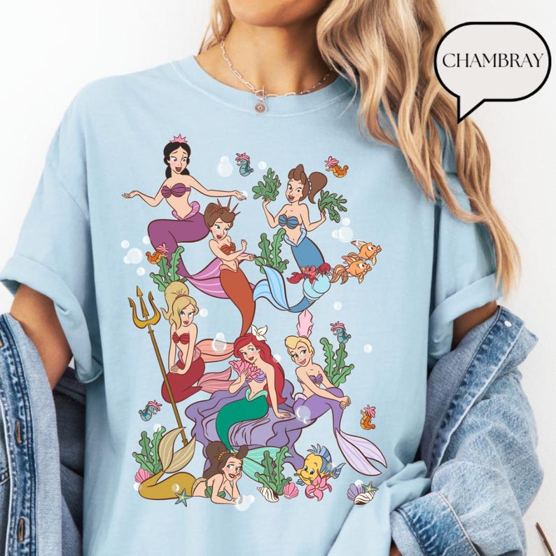 Op de afbeelding: Lichtblauw T-shirt met een kleurrijke afbeelding van zeemeerminnen, vissen en zeewier. Het ontwerp toont diverse zeemeerminnen met verschillende haar- en staartkleuren. Het woord "CHAMBRAY" staat in een tekstballon rechtsboven.