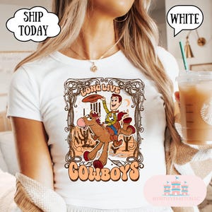 May include: White t-shirt featuring a cartoon cowboy riding a horse, with the words "LONG LIVE COWBOYS" in a vintage style. The design includes a cowboy hat and desert scenery. The shirt has a crew neck.