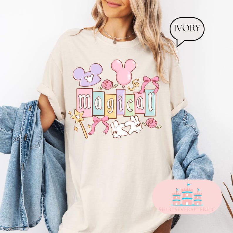 Op de afbeelding: Ivoor kleurig T-shirt met het woord "magical" in pastelkleuren, met Mickey Mouse oren, een ballon, rozen, een toverstaf en een konijn. Het shirt heeft opgerolde mouwen en wordt gedragen met een spijkerjasje.