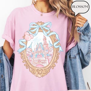 Puede incluir: Camiseta rosa claro con una ilustraci&oacute;n enmarcada en oro de un castillo de cuento de hadas con detalles azules y un lazo azul claro. La palabra "BLOSSOM" est&aacute; en una burbuja. La camiseta es de material suave.