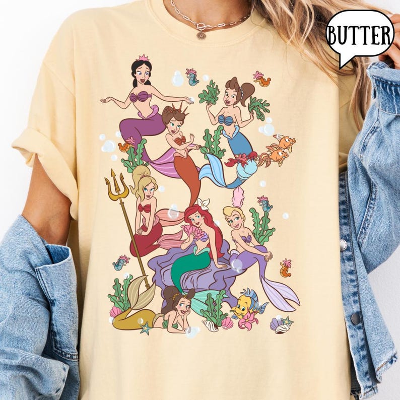 Op de afbeelding: Lichtgeel T-shirt met een kleurrijke illustratie van zeemeerminnen, zeewier en vissen. Het ontwerp toont diverse zeemeerminnen met verschillende haarkleuren en outfits. Het woord "BUTTER" staat in een tekstballon rechtsboven.